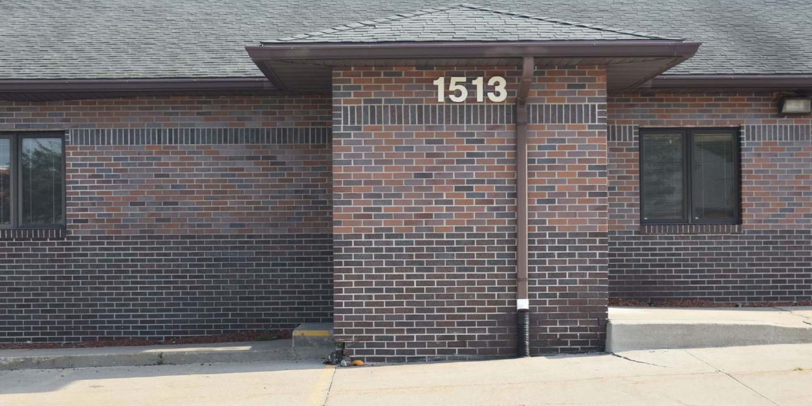A brick building with the number 1513 displayed above a central column, flanked by two dark windows, and a concrete parking lot in front.