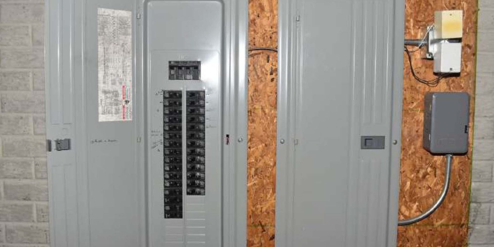 Two gray electrical breaker panels mounted on a wood wall; the left panel door is open showing circuit breakers and a diagram, while the right panel door is closed. Several wires enter the tops of both panels.