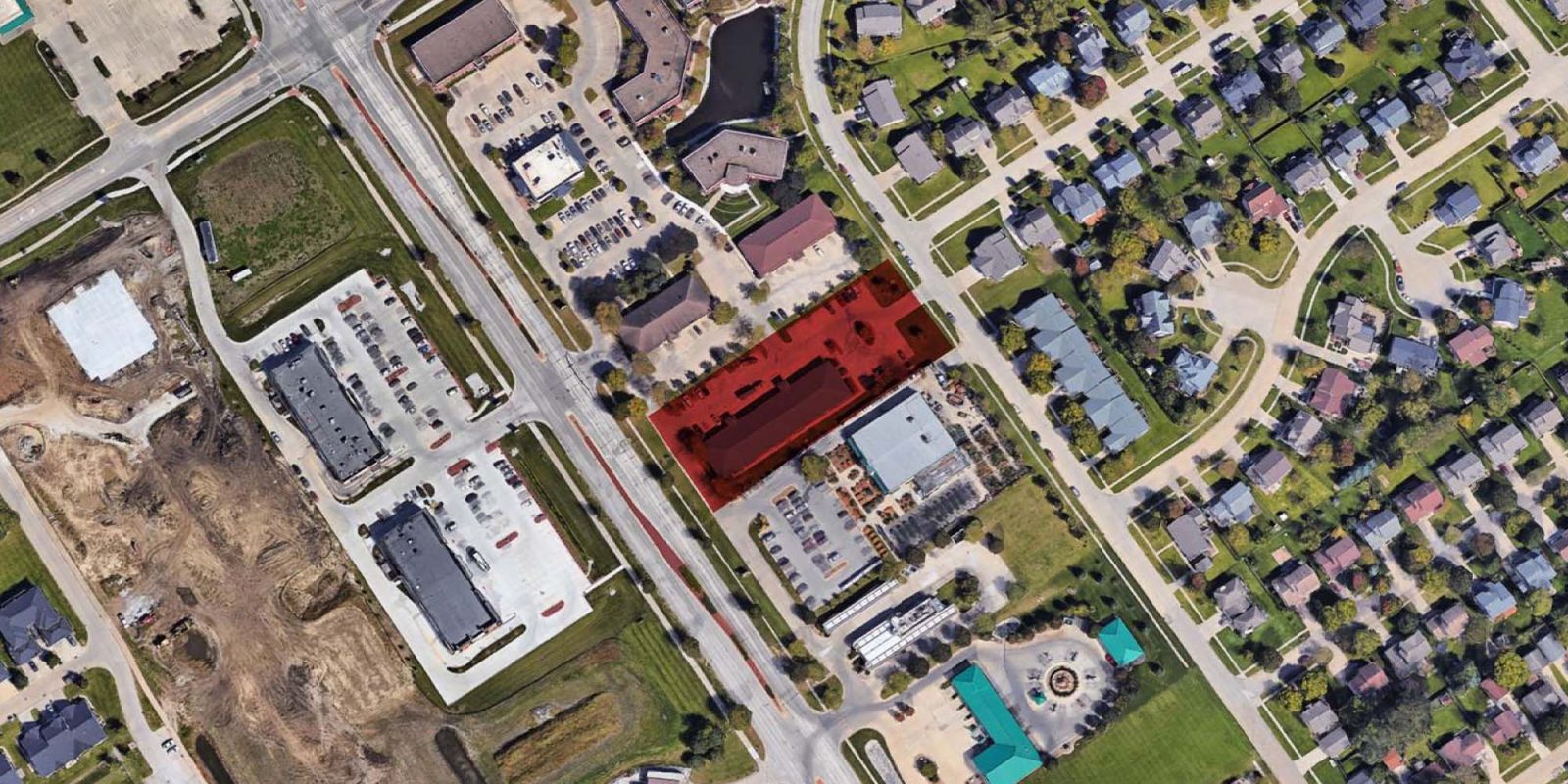 Aerial view of a suburban neighborhood with houses, roads, parking lots, and businesses. A rectangular area in the center is highlighted in red, indicating a specific property or location among nearby buildings and green spaces.
