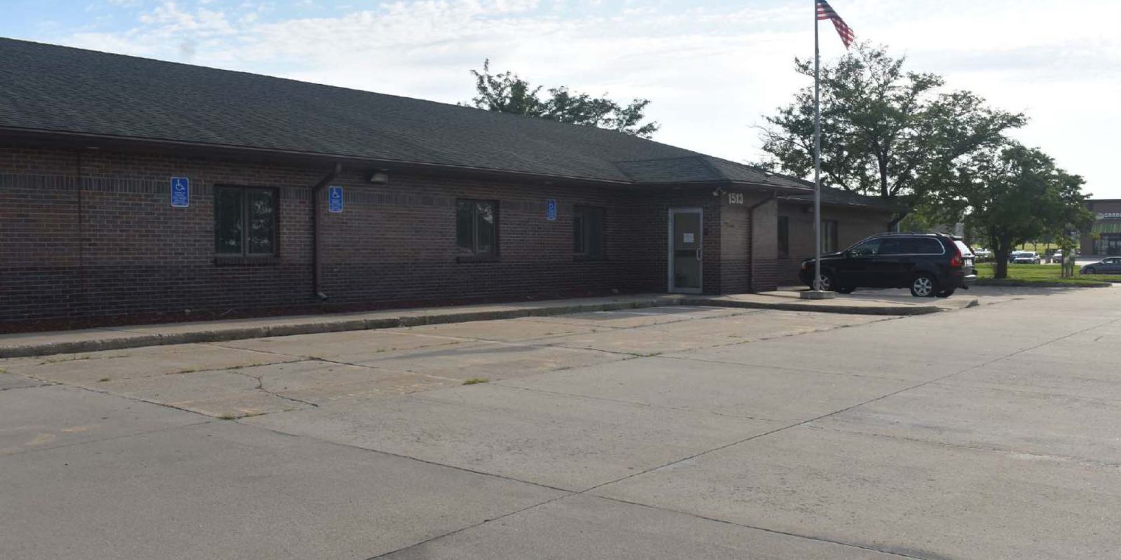 A single-story brick building with several accessible parking spots, a parked SUV near the entrance, and an American flag on a pole in front. The sky is partly cloudy.