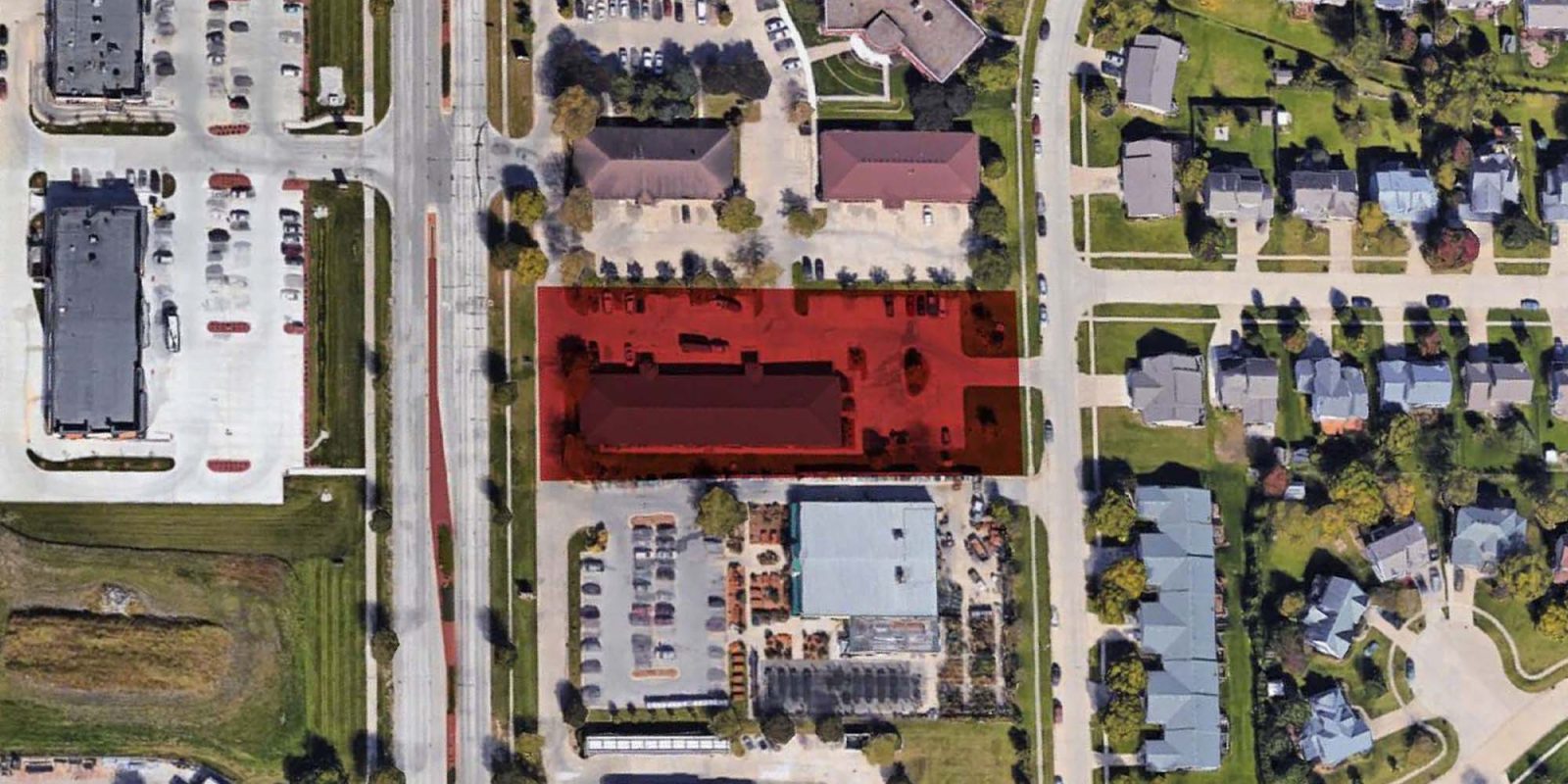 Aerial view of a suburban area with a red highlight over a rectangular building and parking lot, surrounded by roads, businesses, and residential houses with green lawns.