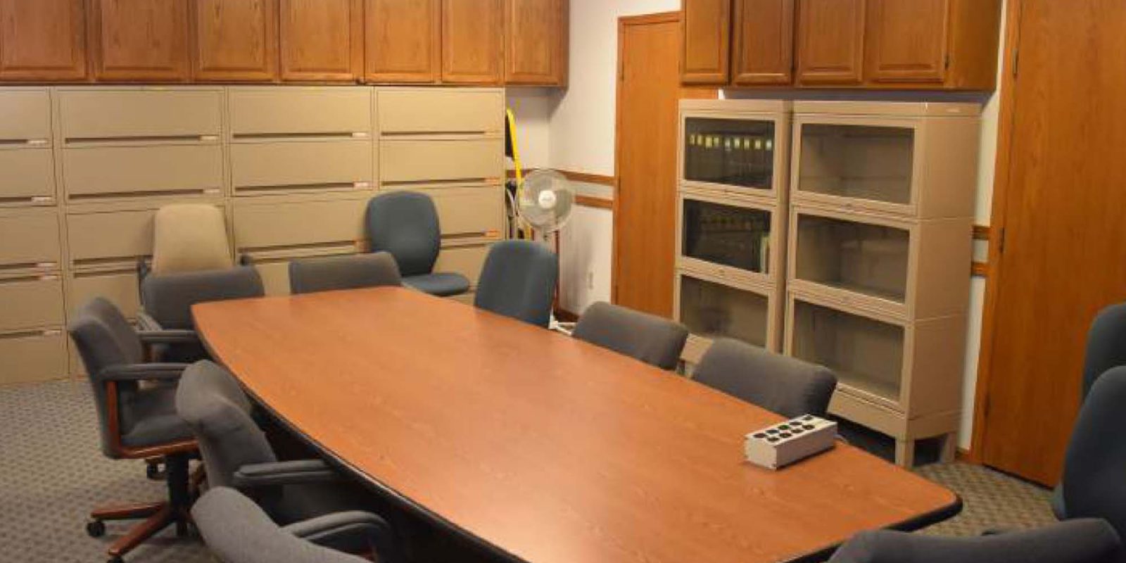 A conference room with a long wooden table surrounded by gray chairs, filing cabinets and wooden cabinets along the walls, a small fan, and a light panel in the ceiling.