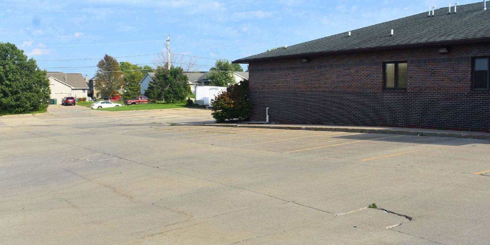 An empty parking lot beside a brick building under a blue sky with scattered clouds; a few cars and trees are visible in the background.