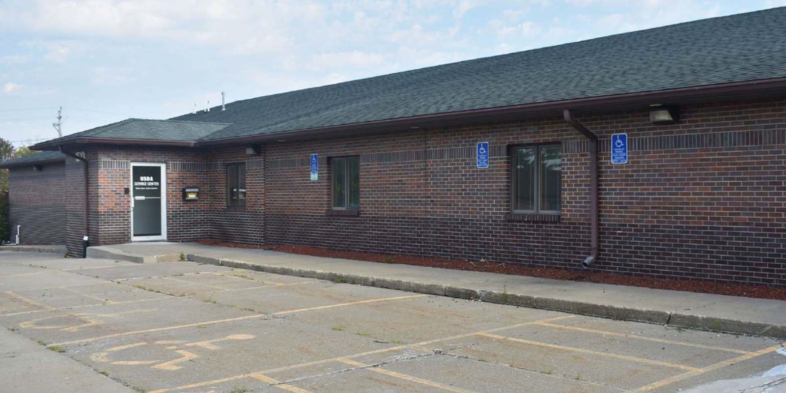 A single-story brick building with a dark roof and several windows. The parking lot in front has marked spaces, including two accessible parking spots near the entrance, and faded "No Parking" text on the pavement.