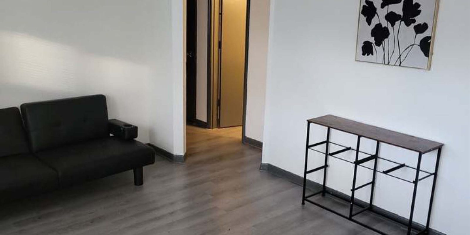 A minimalist room with light gray wood flooring, a black futon sofa on the left, a metal and wood console table on the right, and a black floral artwork on the white wall. An open doorway leads to a lit hallway.