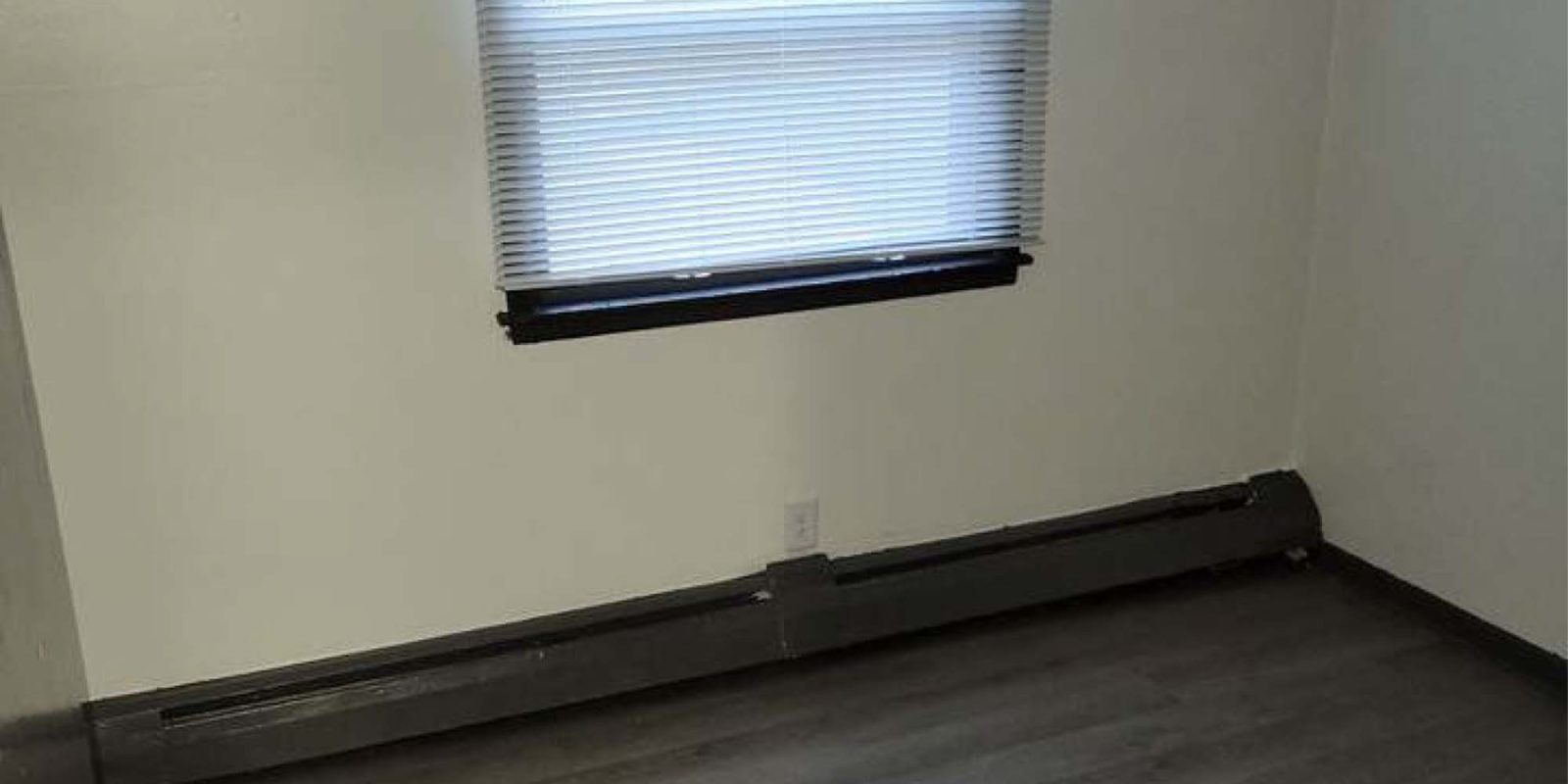 An empty room with white walls, a window with closed blinds, dark trim, gray wood flooring, and a baseboard heater along the wall beneath the window.