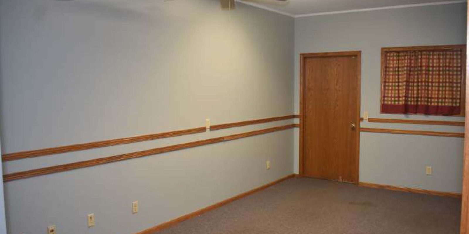 An empty room with light gray walls, brown trim, gray carpet, a wooden door, a small window with a plaid curtain, a ceiling fan, fluorescent lighting, and multiple electrical outlets along the walls.