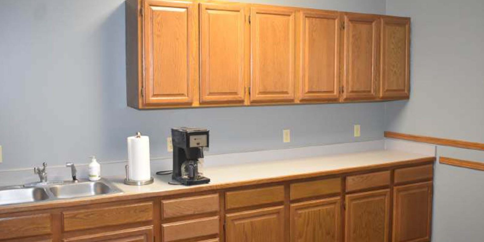 A simple kitchen with wooden cabinets, a white countertop, a stainless steel sink, a paper towel holder, a coffee maker, and a soap dispenser against a gray wall.