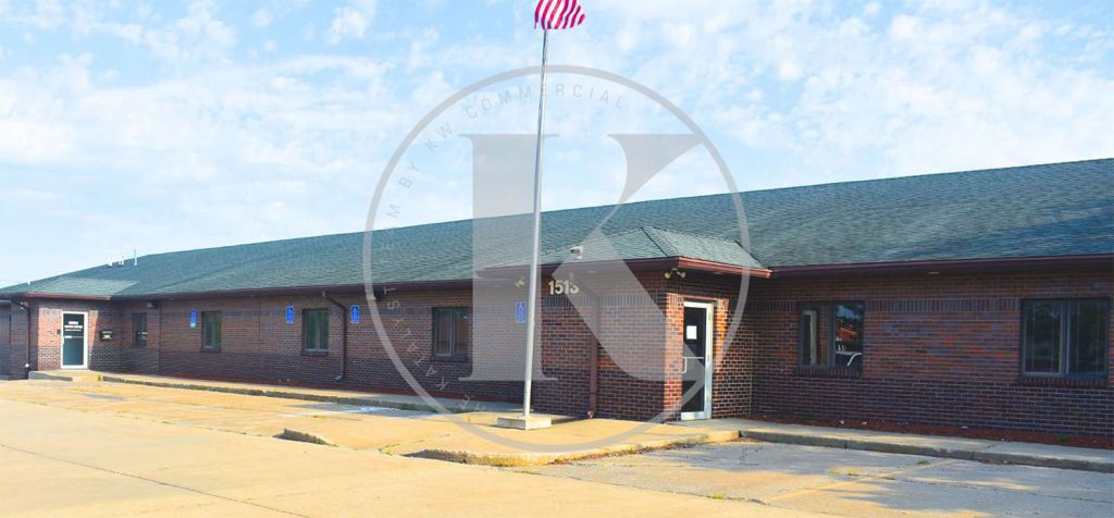 A single-story brick commercial building with a dark roof, several windows, and a central entrance. An American flag is flying above the entrance. The parking area in front is empty under a partly cloudy sky.