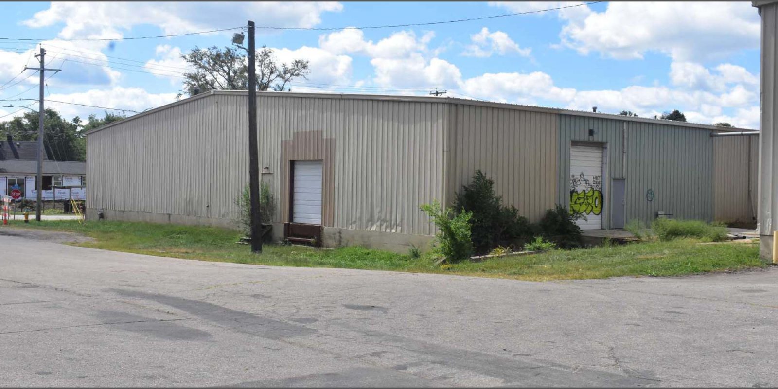 A beige metal warehouse with a closed white garage door and graffiti on one entry door sits at a street corner, with grass and weeds growing along its base under a partly cloudy sky.