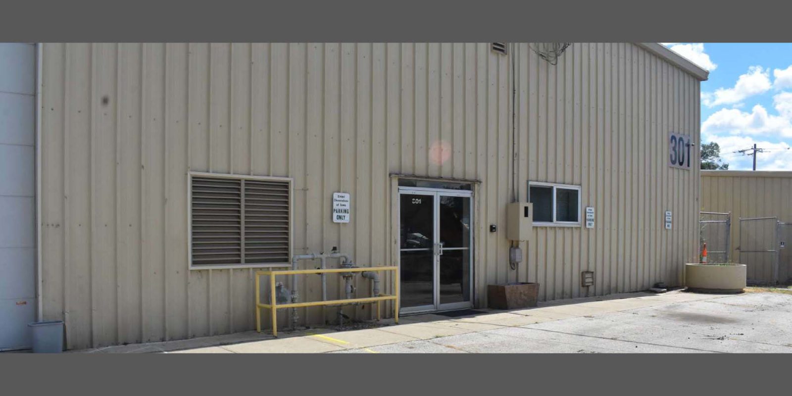 A beige industrial building with a glass door marked 204, two small windows, yellow railings, and signages on the wall. The number 301 is visible on the upper right. The sky is partly cloudy.