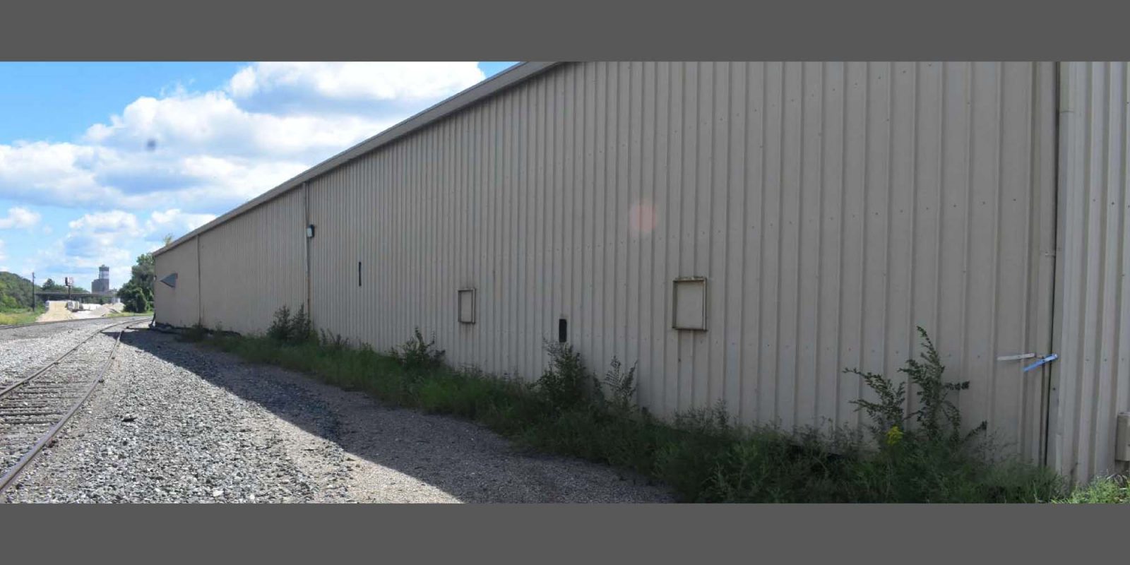 A beige metal warehouse wall runs alongside a gravel path with railroad tracks on the left. Weeds grow at the building's base. The sky is blue with scattered clouds.