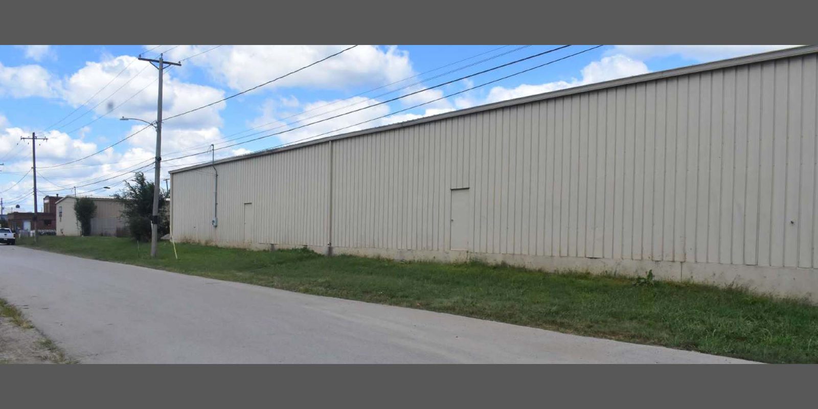 A long, beige industrial warehouse with metal siding sits beside a quiet, empty street under a partly cloudy sky. Grass lines the side of the building, and utility poles run along the road.