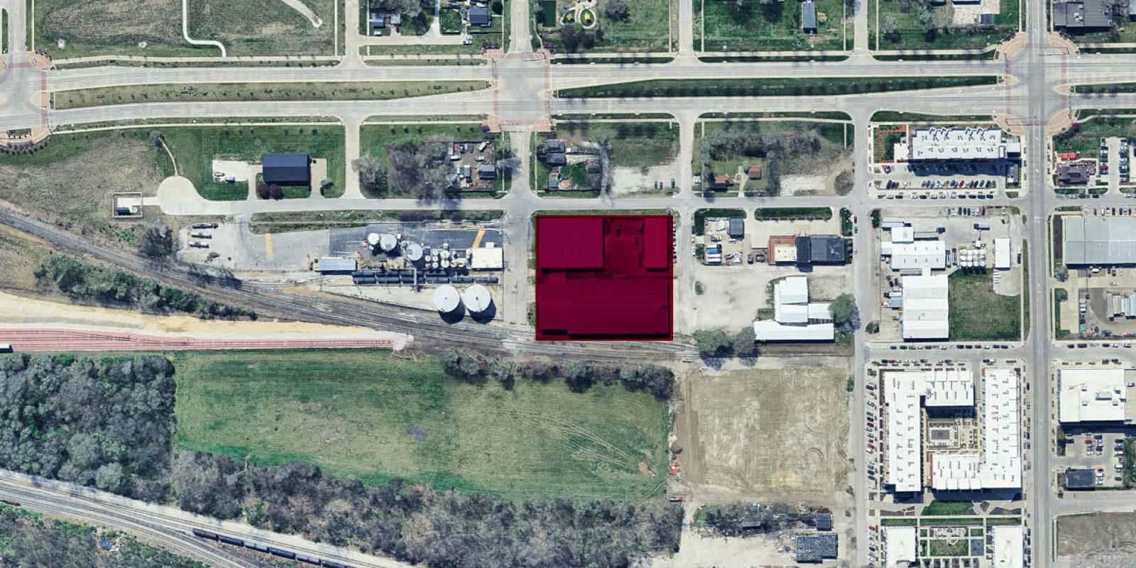 Aerial view of an urban area featuring buildings, roads, and green spaces. A group of buildings near the center is highlighted in dark red, surrounded by industrial and commercial properties.