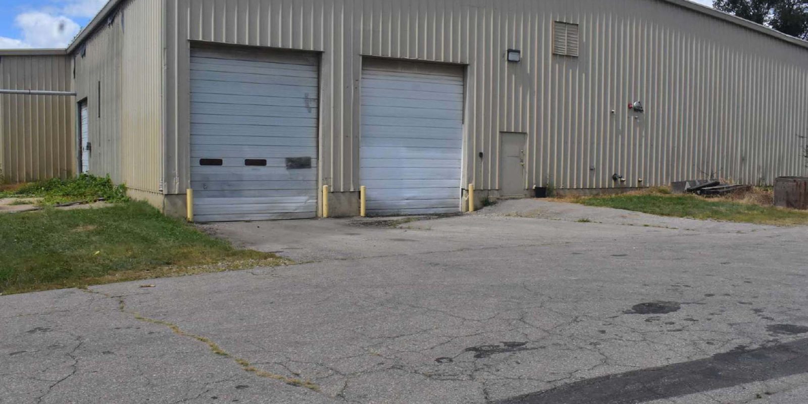 A large beige industrial warehouse with two closed garage doors, surrounded by cracked pavement and patches of grass under a partly cloudy sky.