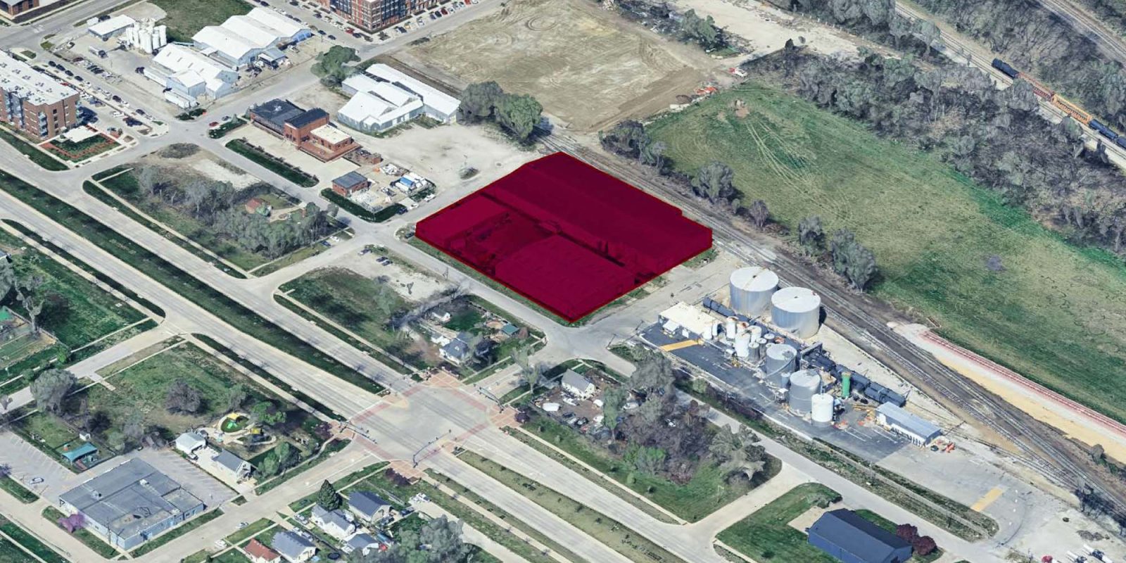 Aerial view of an urban area with streets, buildings, trees, and an industrial site. A rectangular property near the center is highlighted in red. Residential and commercial buildings surround the marked area.