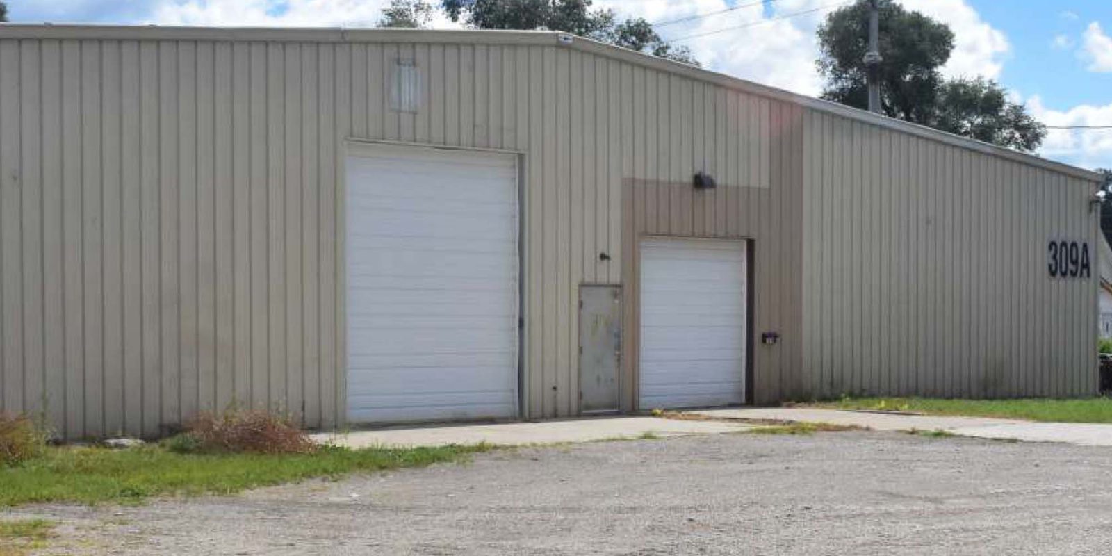 A beige metal warehouse with two large garage doors and a smaller door, situated on a gravel lot under a partly cloudy sky. The building displays the number 309A on the right side.