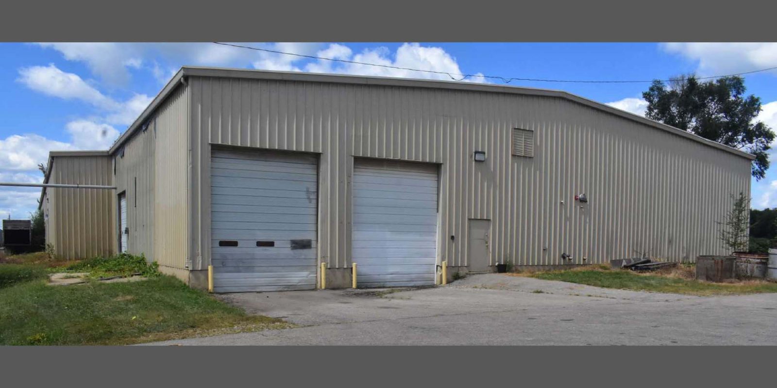 A beige industrial warehouse with two large garage doors, a smaller entry door, and a paved driveway. The building sits on a grassy area under a partly cloudy sky.