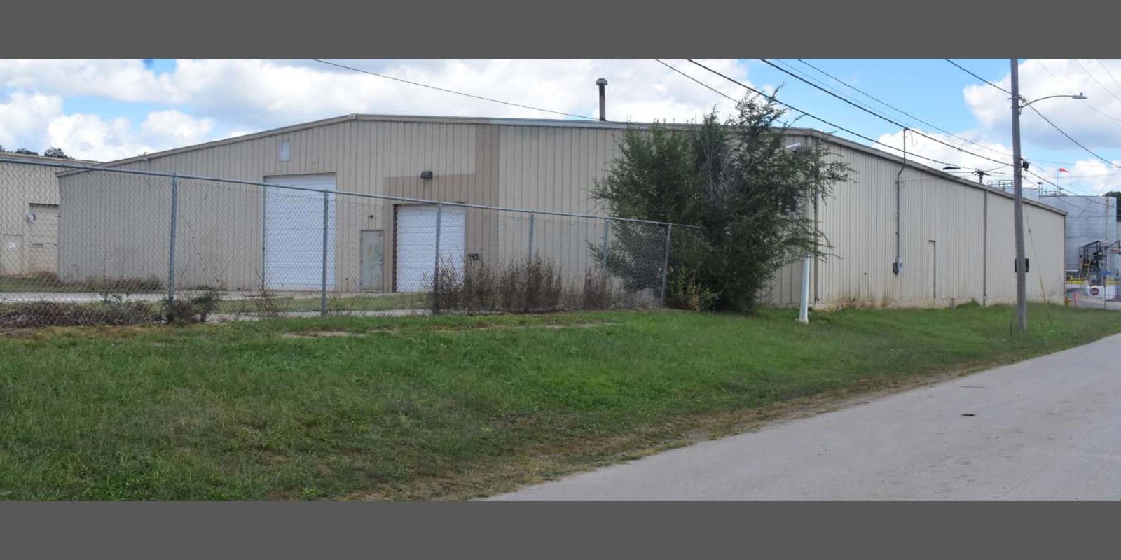 A beige industrial warehouse with a metal fence and overgrown grass in front, situated beside a paved road under a partly cloudy sky.