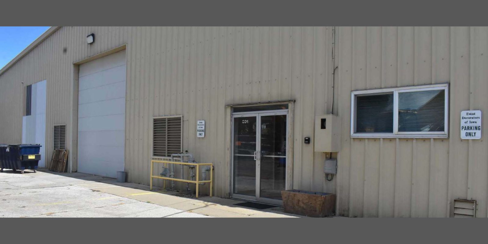 A beige industrial warehouse with a large garage door, metal siding, glass entry door, small window, and a "Reserved Customer Parking Only" sign on the wall. A trash bin and yellow railings are near the entrance.