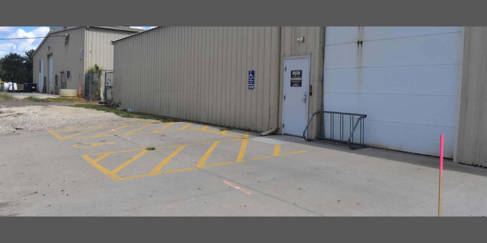 A beige industrial building with a large white garage door, an accessible entrance door with a sign, yellow diagonal lines marking an accessible parking zone, and a bike rack on a concrete surface.