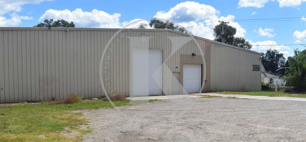 A beige industrial warehouse with two white garage doors and a small entry door, marked “3031,” sits on a gravel lot under a partly cloudy sky. Sparse grass and weeds grow along the building’s edge.