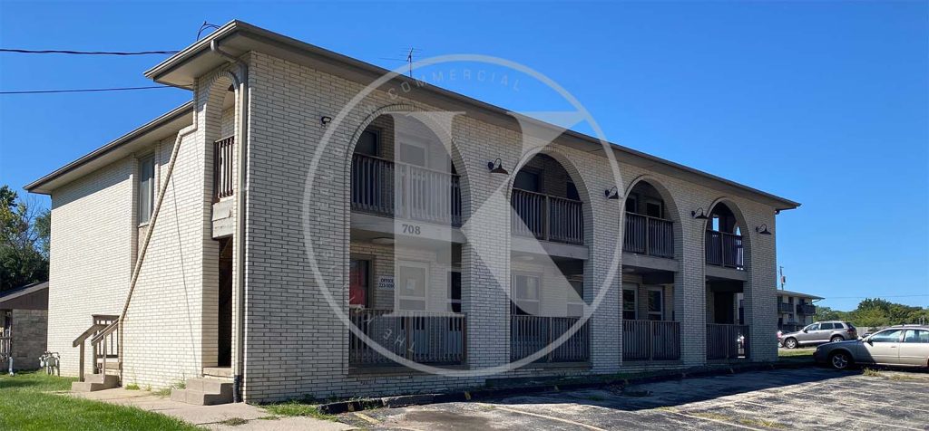 Two-story brick apartment building with arched openings and wooden railings on the upper level, set against a clear blue sky with a grassy area and paved parking spaces in front.