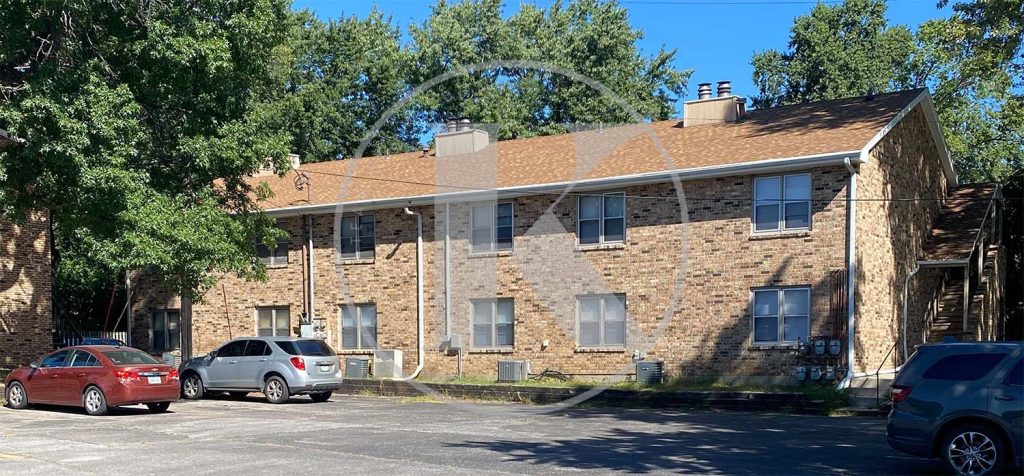 A two-story brick apartment building with a brown roof, surrounded by trees, and several parked cars in front on a sunny day.