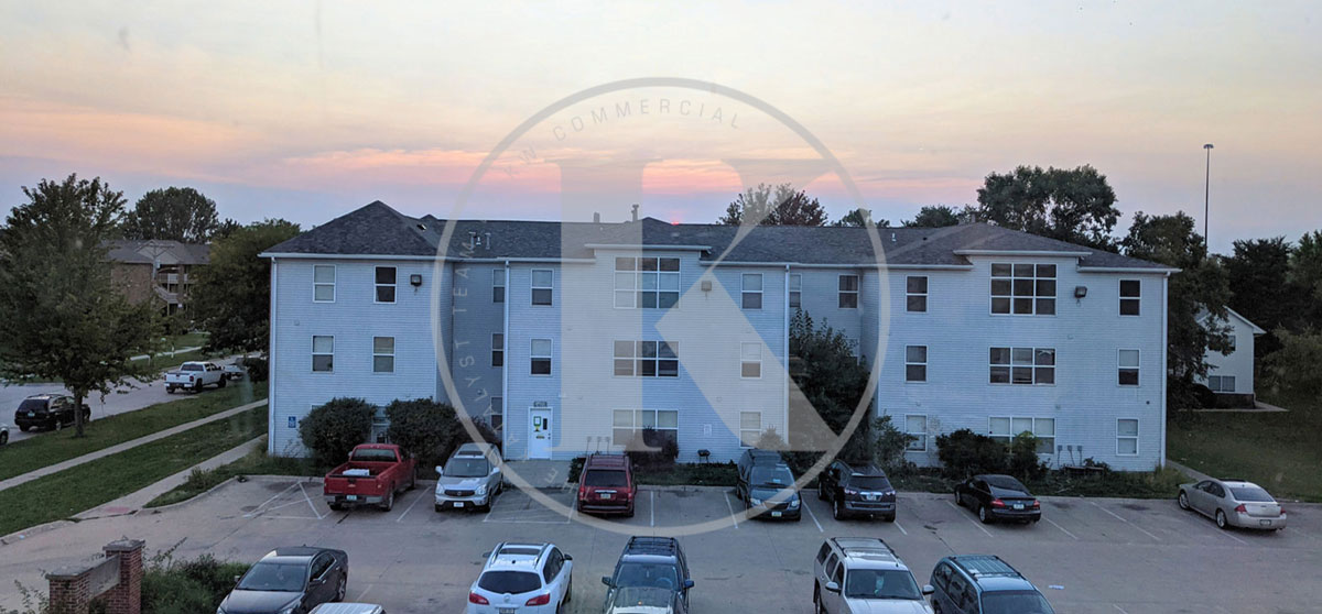 A three-story light blue apartment building with multiple windows, surrounded by trees and a parked row of cars in the parking lot at sunset. The sky has a soft pink hue.