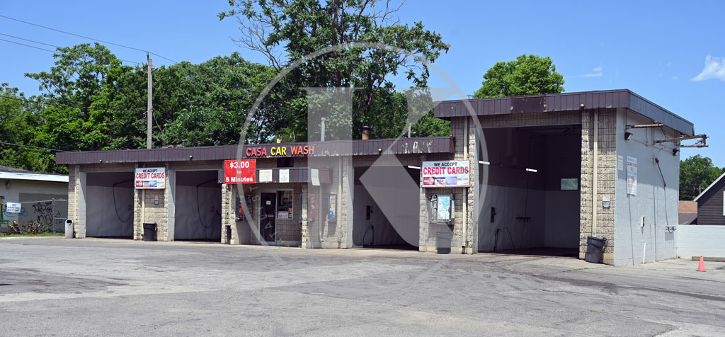 A self-service car wash with four open bays, signs indicating the price, and instructions on the front. The building is made of concrete blocks and sits on an empty paved lot with trees and a blue sky in the background.