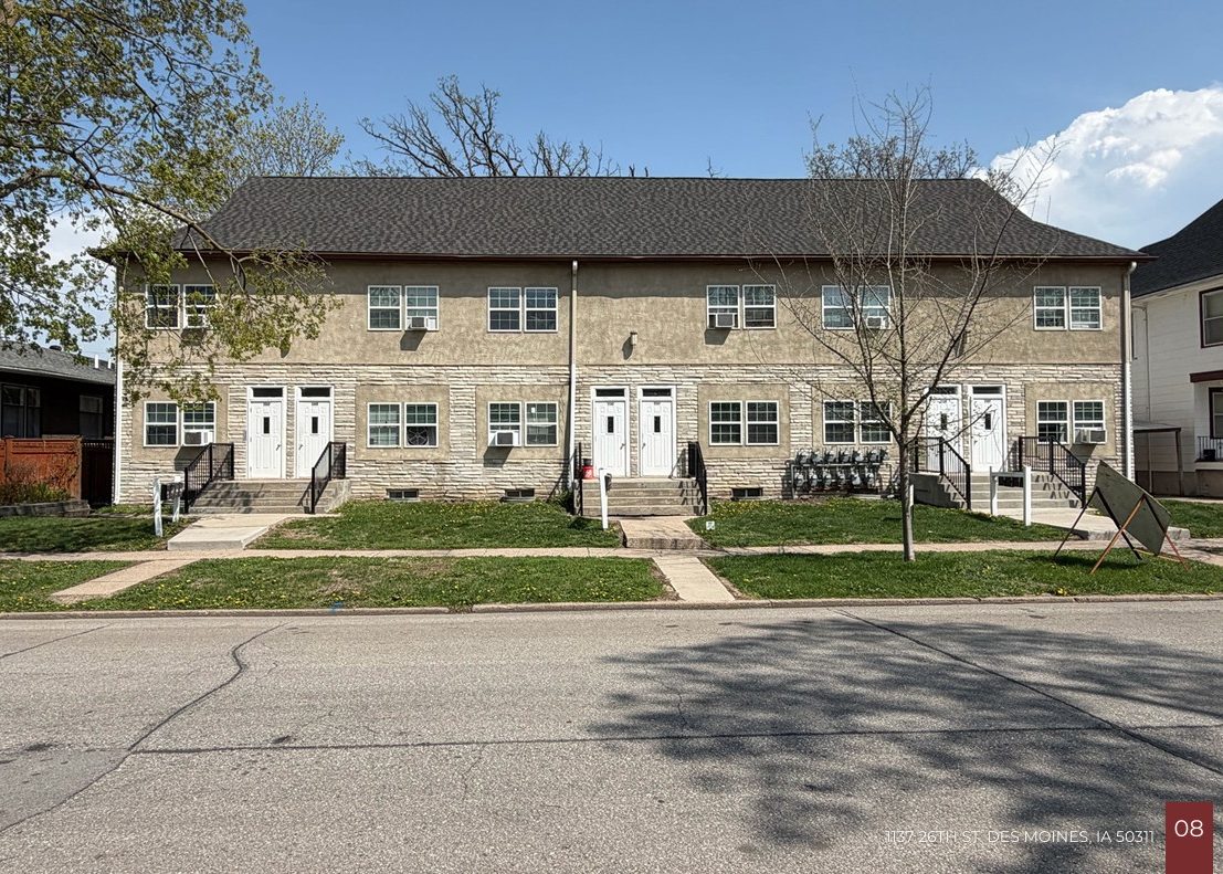 A beige two-story apartment building with four white doors, steps leading up to each entrance, small front lawns, and leafless trees along the sidewalk under a clear sky.