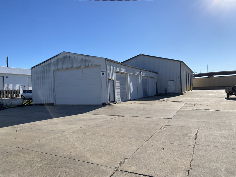 Two commercial industrial buildings labeled B and C with multiple roll-up garage doors, situated on a large concrete lot under a clear blue sky.