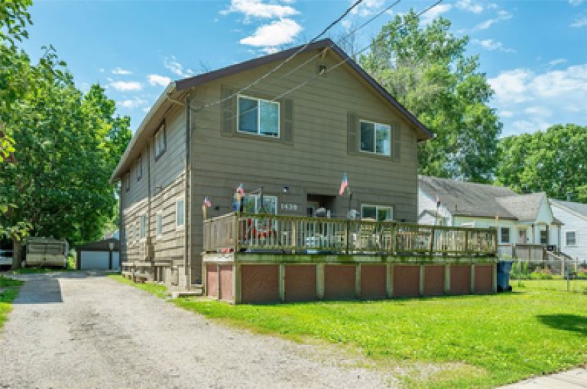 A two-story house with brown siding and a large wooden deck, featuring a gravel driveway and a grassy front yard.