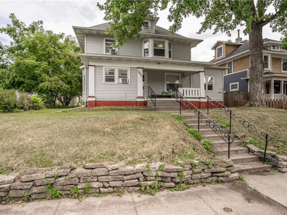 Two-story gray house with a front porch, stone retaining wall, and concrete steps leading to the entrance.