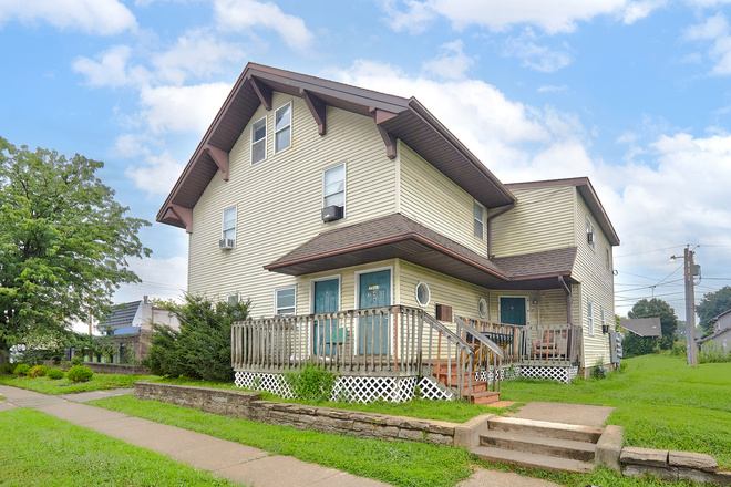 Two-story beige house with a wrap-around wooden deck and multiple entrances, viewed from the street.